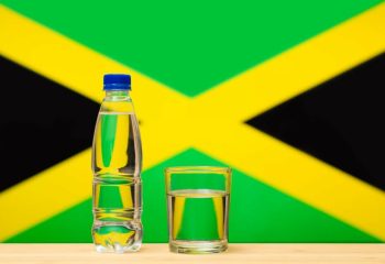 A bottle of clean drinking water and a glass stand on the table against the background of the flag of Jamaica. A concept for the supply of clean drinking water in Jamaica.